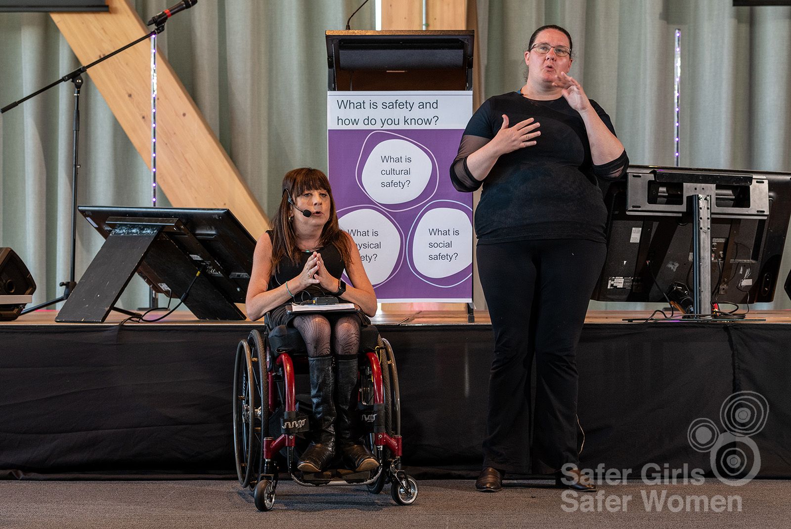 Mel Harrison sits in her wheelchair delivering a speech at the Safer Girls Safer Women 2022 symposium, next to an Auslan interpreter signing.