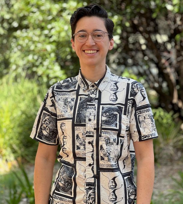 Millie stands smiling in front of greenery. They have a very cool comic book print on their button-down shirt, and they look generally awesome (and also wrote this alt text).