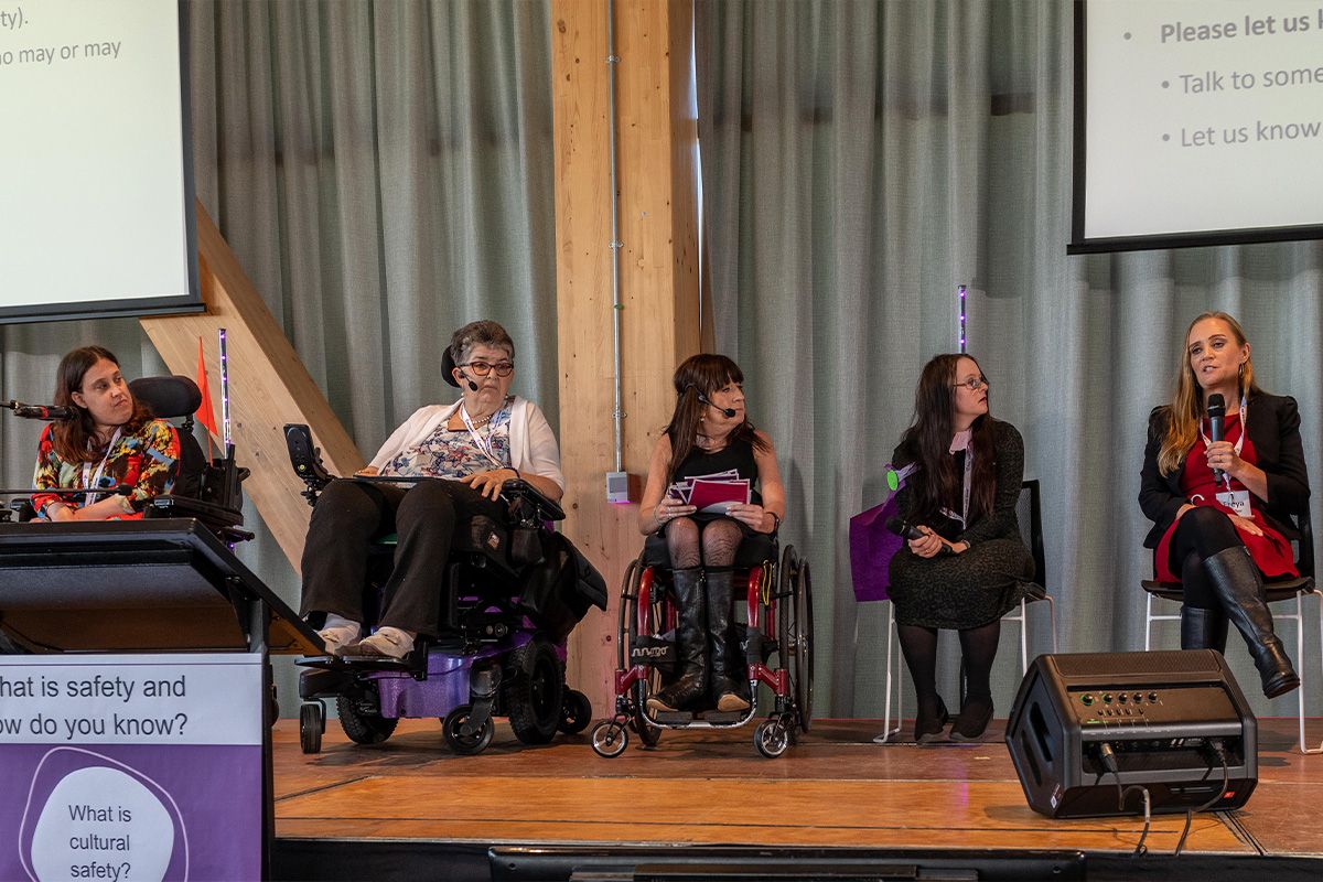 A panel of speakers using chairs and wheelchairs speak on stage at the 2022 Safer Girls Safer Women symposium.