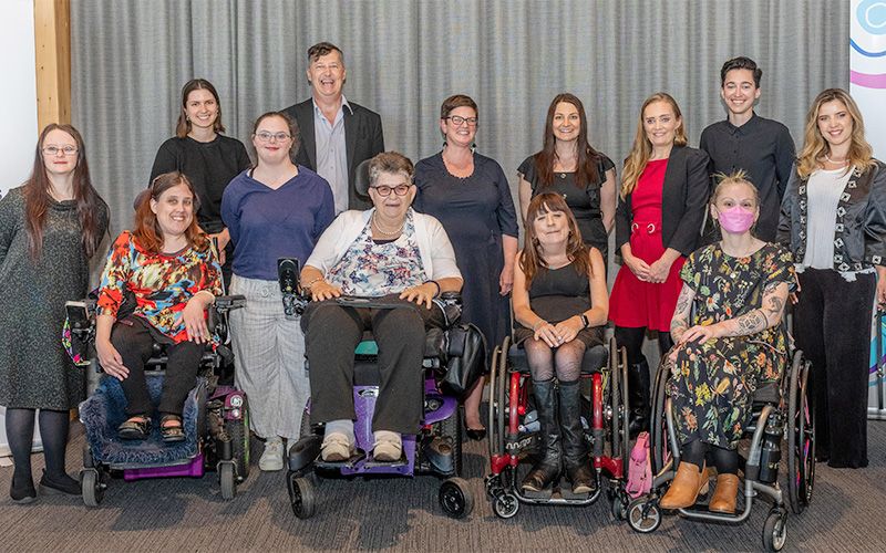 Photo of 13 people standing or using wheelchairs, smiling for a group photo at the 2022 Safer Girls Safer Women symposium.