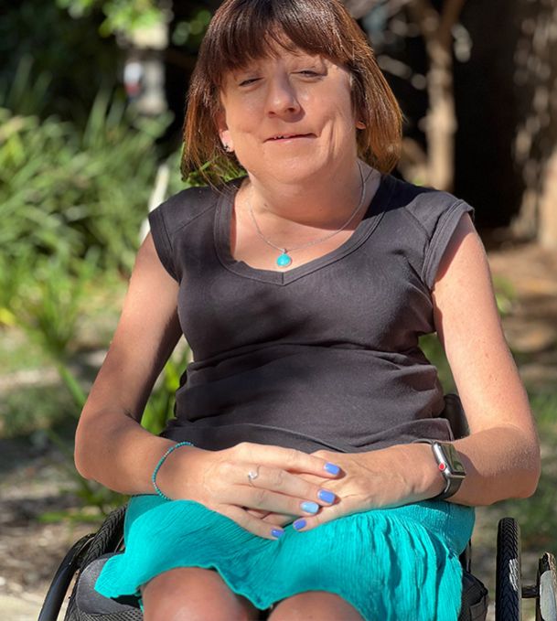 Mel sits smiling in her wheelchair in front of some nature in the background. She has paired her trademark blue nail polish with an aqua skirt (and necklace).